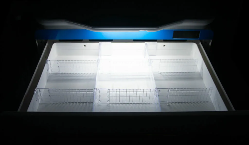 Open freezer chest with blue trim and white wire shelving baskets inside, brightly lit interior against black background.