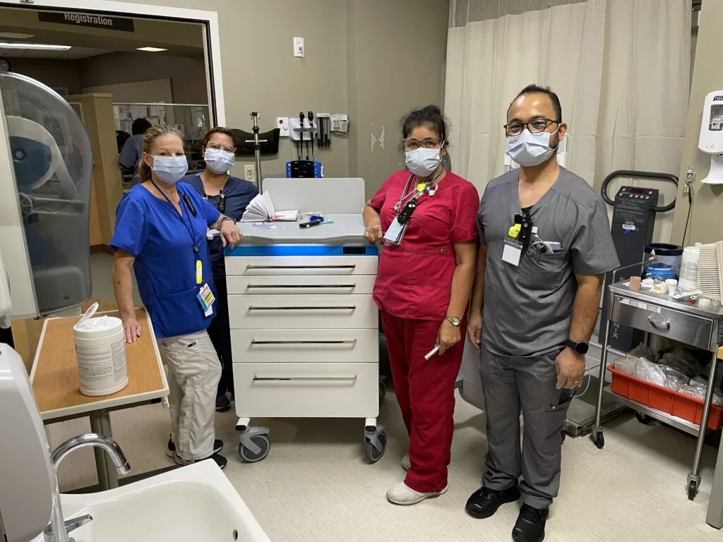 Four healthcare workers wearing masks stand in a medical facility next to a white mobile cart with blue top.