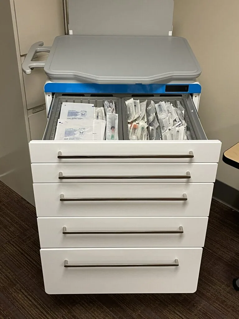 Medical supply cart with five white drawers and blue top, top drawer open displaying medical documents and packets