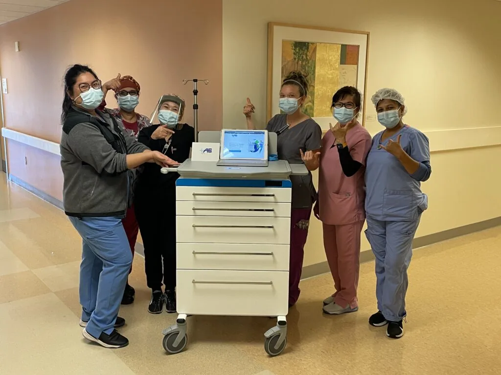 Six healthcare workers wearing masks posing around a medical cart with a laptop in a hospital corridor