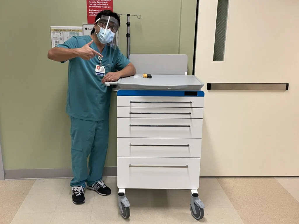 Healthcare worker in teal scrubs and mask standing beside white medical supply cart with multiple drawers and blue top