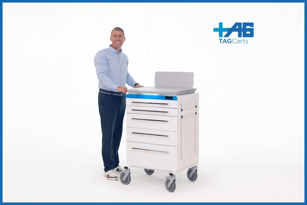 Man in light blue shirt standing beside white medical cart with blue top and multiple drawers on casters