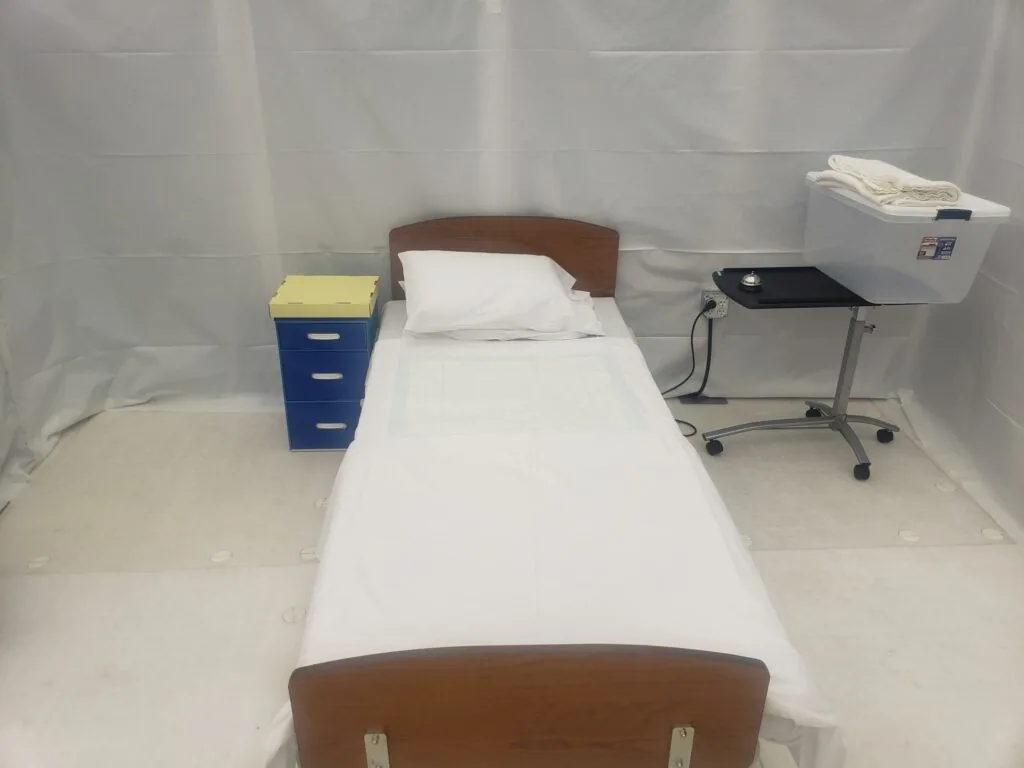 Single bed with white linens and wooden headboard in a room with white curtain walls, blue nightstand on left and medical equipment on right