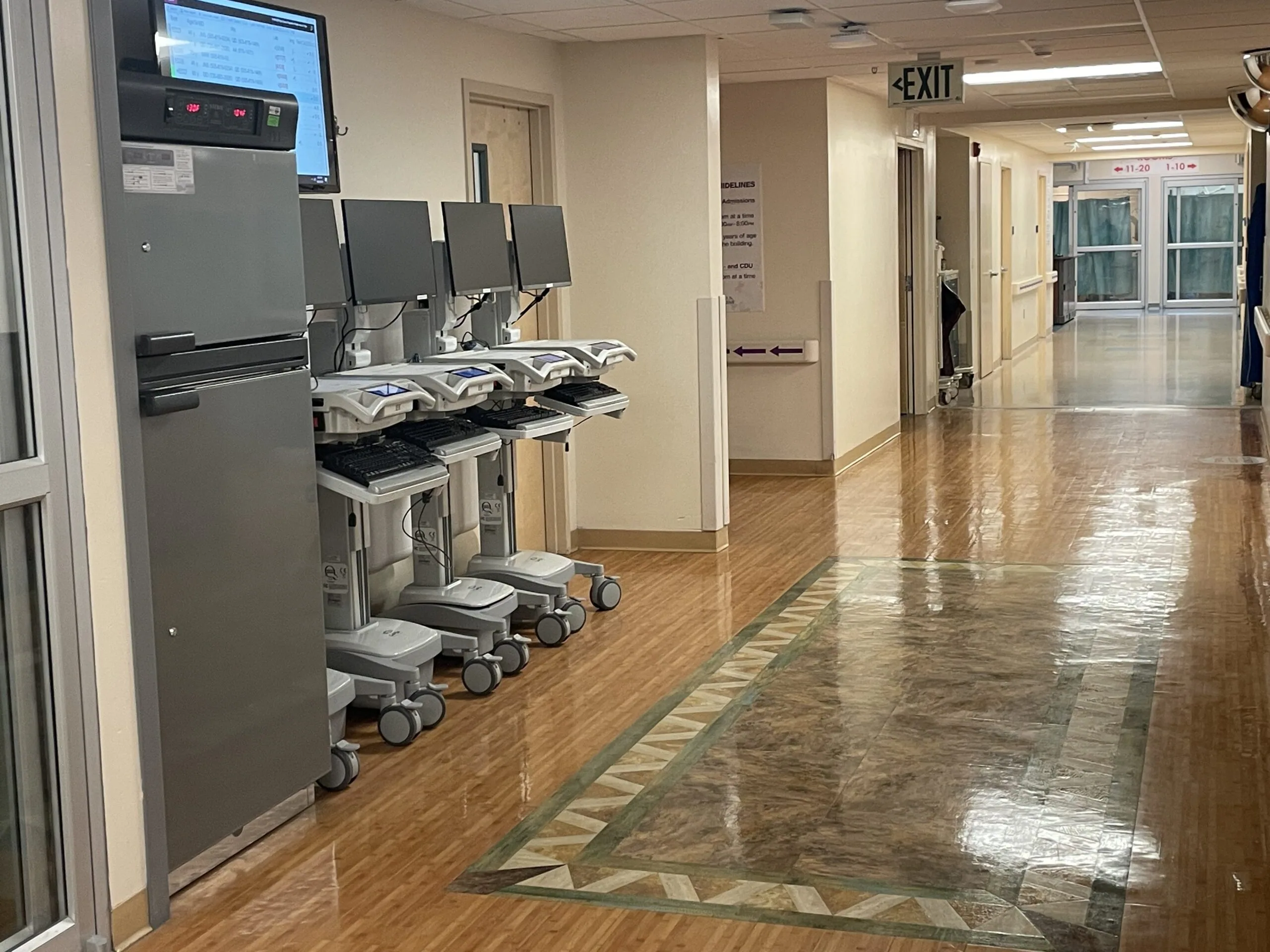 Hospital corridor with medical equipment carts lined against wall, exit sign visible in distance