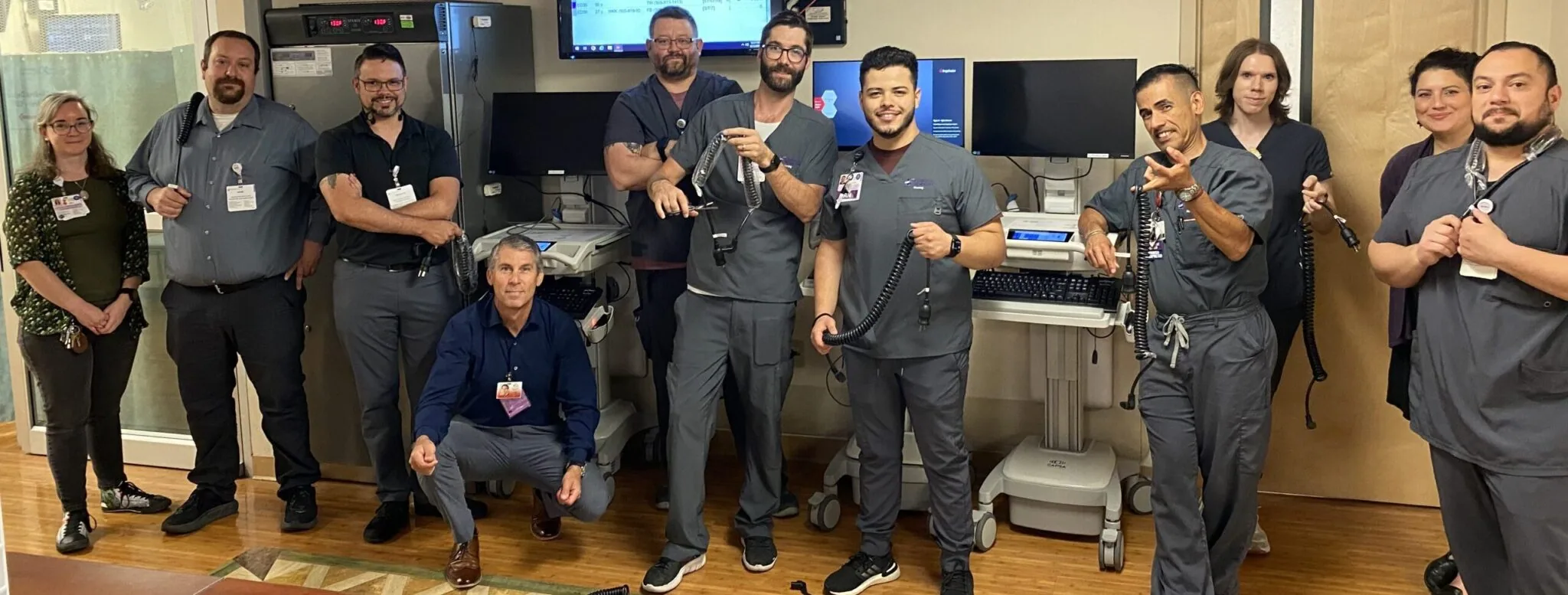 Group of medical or technical staff in dark uniforms posing in a clinical or laboratory room with equipment and monitors in background