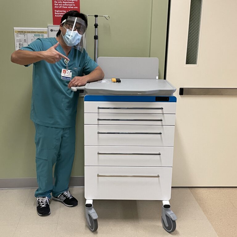 Healthcare worker in teal scrubs and mask standing beside white medical supply cart with multiple drawers and blue top