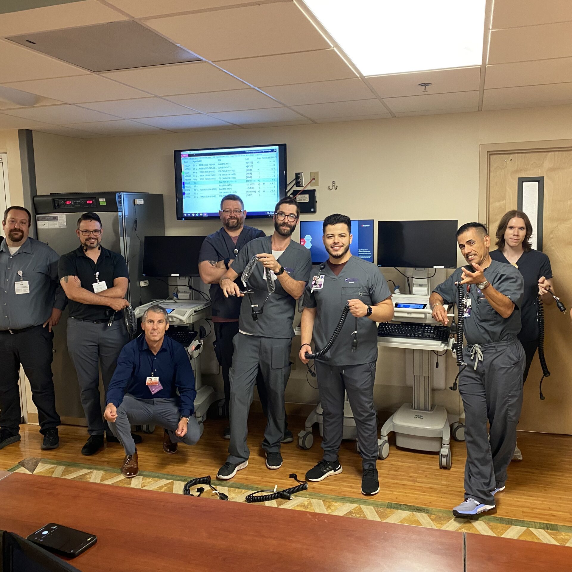 Group of medical professionals in gray scrubs posing together in a hospital break room with medical equipment and monitors visible